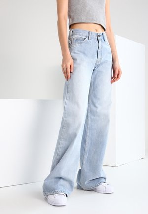 Personne portant un jean large bleu clair, un débardeur gris court et des baskets blanches, debout devant un fond blanc uni.