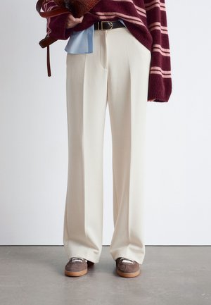 Cream wide-leg trousers with a textured fabric, paired with a maroon striped sweater and brown shoes with white laces, standing on a gray surface.