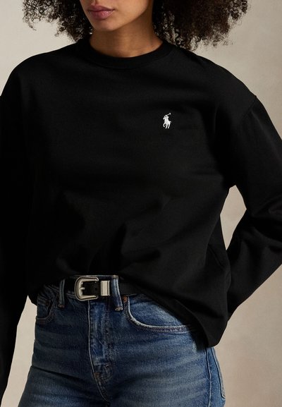 Kobieta nosząca czarną koszulę z długim rękawem z małym, białym logo polo, włożoną w wysokie niebieskie jeansy z czarnym paskiem.