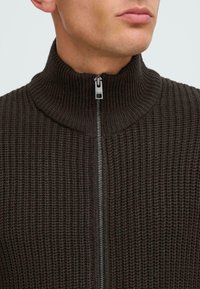 Brauner gestrickter Reißverschluss-Pullover mit hohem Kragen, der ein strukturiertes Muster und einen silbernen Reißverschluss vorne in der Mitte aufweist.