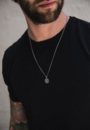 Silver pendant necklace hangs on a thin chain, worn over a fitted black t-shirt. Tattoo visible on the forearm, background is neutral.
