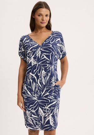 Kobieta ubrana w granatową sukienkę do kolan z białym botanicznym wzorem, dekoltem w serek, krótkimi rękawami, trzymająca ręce w bocznych kieszeniach, stojąca na jednolitym tle.
