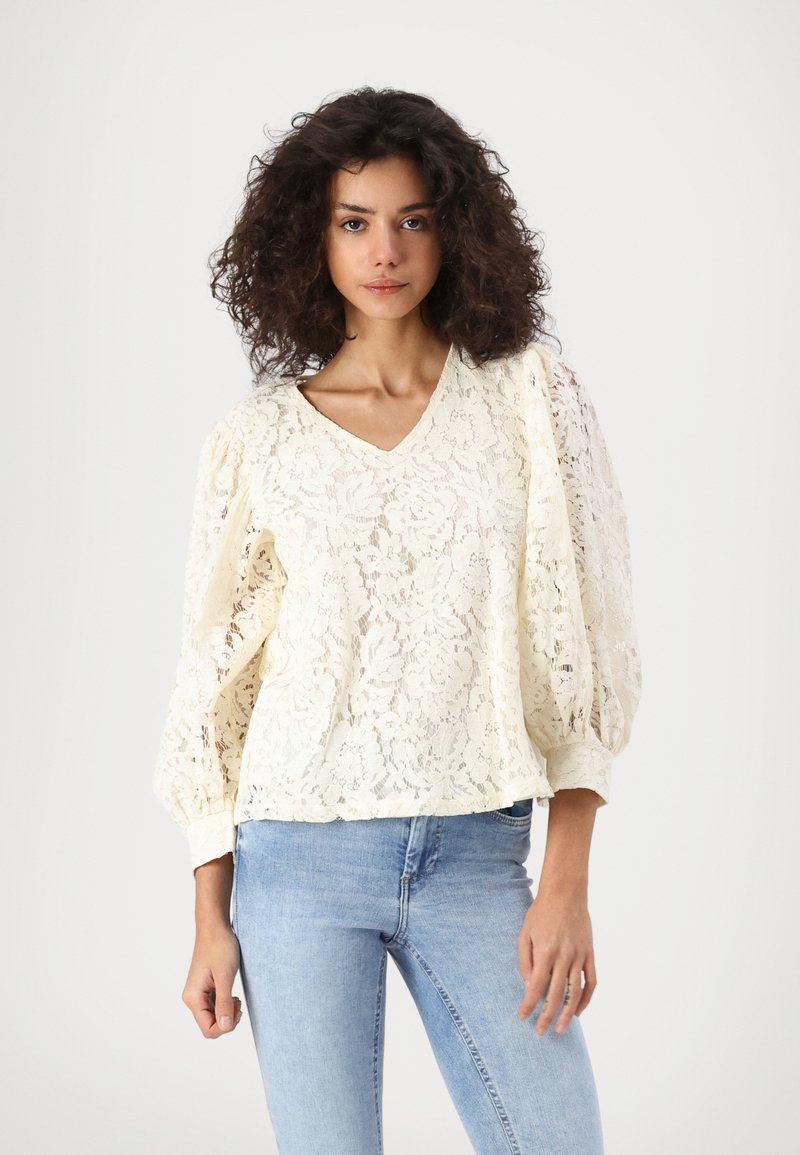 Blusa in pizzo color crema con scollatura a V, maniche a sbuffo e motivo floreale. Abbinata a jeans blu, mette in evidenza un look rilassato e un design testurizzato.