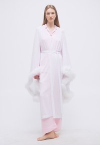 Γυναίκα που στέκεται ξυπόλητη, φοράει ένα ανοιχτό ροζ ρόμπα με αφράτη λευκή επένδυση στα φαρδιά μανίκια και αντίστοιχο παντελόνι πιτζάμας.