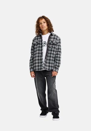 Junge Person mit lockigem Haar, die ein schwarz-weiß kariertes Hemd über einem weißen T-Shirt mit Grafik, dunkle Jeans und schwarze Sneakers trägt und nach vorne schaut.