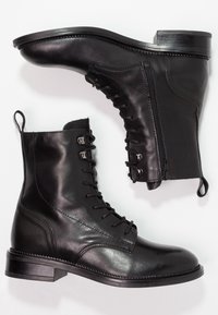 Bottines en cuir noir avec un bout arrondi, un laçage à l'avant, une fermeture éclair sur le côté et un petit talon. Texture lisse avec des détails de coutures.