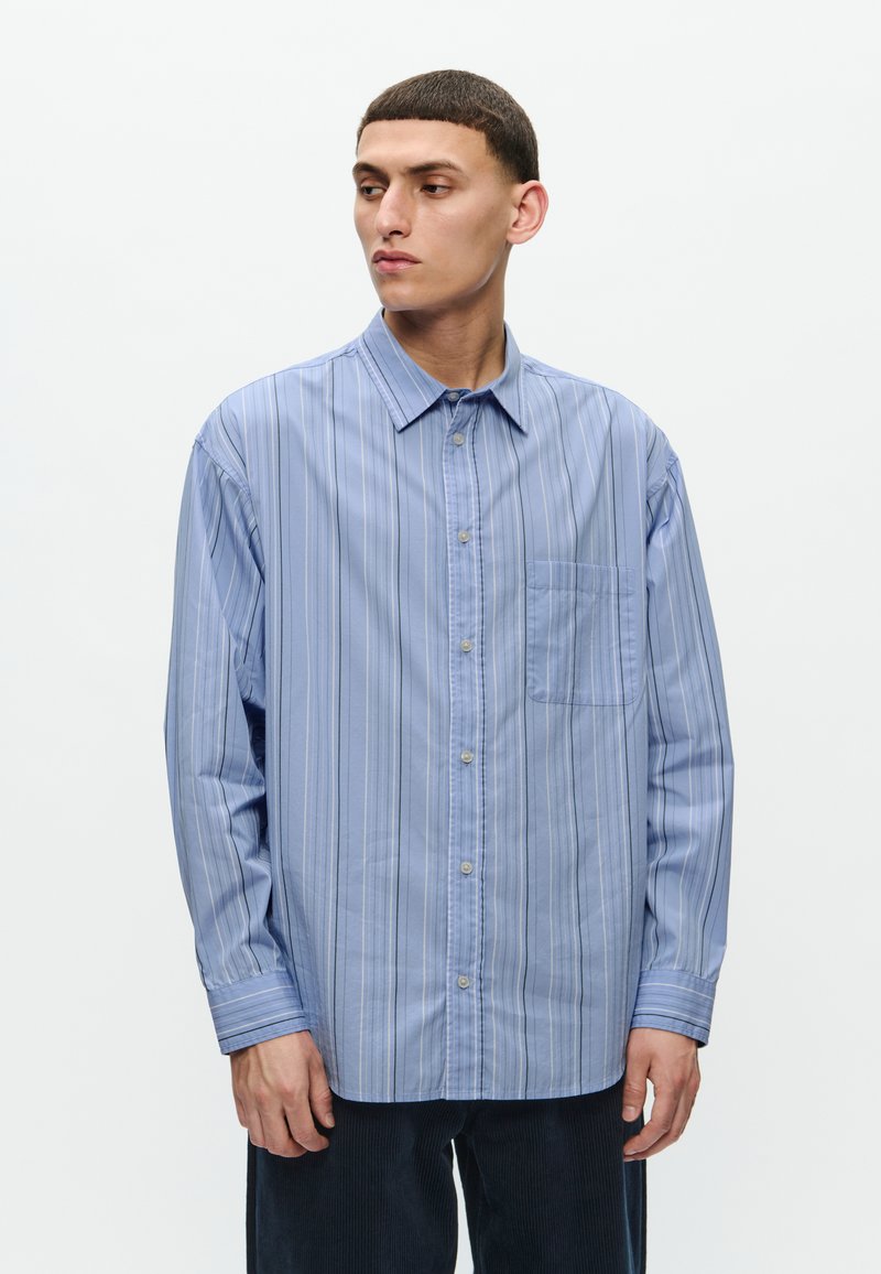 Camisa de manga larga azul claro con rayas verticales, cuello con botones, bolsillo en el pecho y corte relajado. Textura de tela suave.