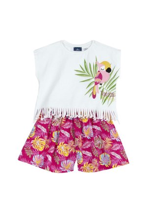 Top in cotone bianco con frange e stampa di pappagallo tropicale, abbinato a pantaloni corti rosa con motivi di foglie colorate.