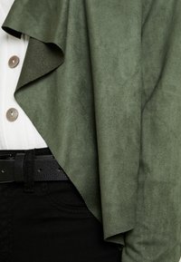 Jemně semišová bunda olivově zelené barvy přes bílou blůzu, doplněná černým opaskem a tmav�ými kalhotami, s hladkou texturou a vrstveným designem.