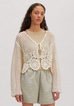 Vrouw draagt een beige mouwloze top en lichtgrijze korte broek, gecombineerd met een losse, witte gehaakte cardigan die vooraan is gestrikt.