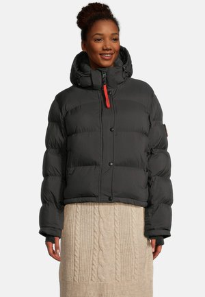 Frau trägt eine schwarze Steppjacke mit Kapuze und Daumenlöchern, kombiniert mit einem beigefarbenen Kabelstrickrock, lächelt und steht vor einem weißen Hintergrund.