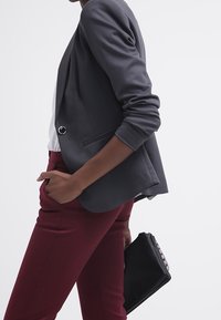 Šedý blejzr s jedním předním knoflíkem a strukturovanými rameny, spárovaný s vínovými kalhotami. Černá kabelka s texturovanými detaily.