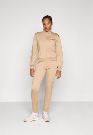 Beige athletic tracksuit featuring a zip-up jacket with a high collar and cuffed sleeves, paired with tapered leggings. White logo accents.