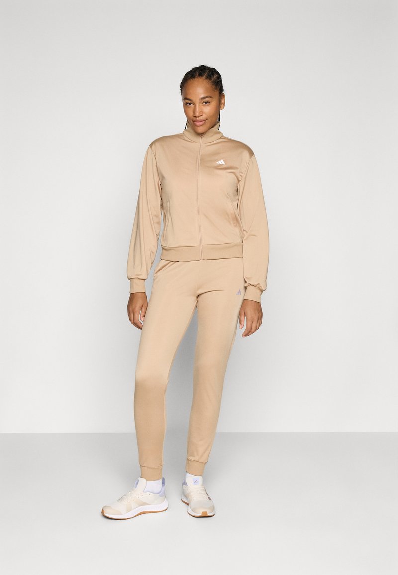 Beige atletisk träningsoverall med en zip-up jacka med hög krage och manschetter, i kombination med tapered leggings. Vita logotyp-accenter.