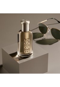 Botella de cristal de colonias BOSS con tapa plateada sobre un pedestal beige junto a unas gafas de sol con montura dorada que proyectan sombras sobre una superficie beige.