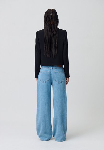 Blazer noir court avec épaulettes, associé à un jean bleu clair à larges jambes comportant cinq poches et une étiquette en cuir brun sur la taille.