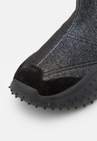 Gros plan sur la pointe d'une chaussure noire avec tissu brillant texturé, détail en daim, et une semelle en caoutchouc avec de petits crampons ronds.