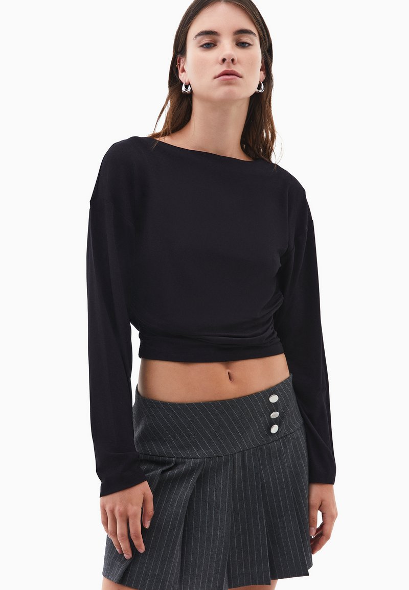 Kvinna som bär en långärmad svart kort topp och en grå randig plisserad kjol med tre vita knappar i midjan.