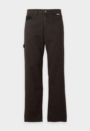 Junger Mann steht mit der Hand in der Tasche, trägt einen schwarz-grauen Iceberg-Pullover mit Hasenmotiv, dunkle Hose und schwarze Sneakers.
