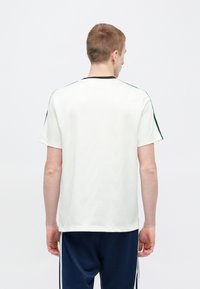 Valge lühikese varrukaga t-särk tekstuurse mustriga, must kaelus ja rohelised detailid õlgadel, vaadatuna tagantpoolt.