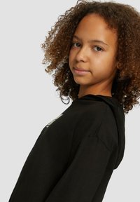 Felpa nera con stampa grafica frontale, realizzata in materiale morbido. Presenta una vestibilità rilassata e un cappuccio con cordino. I capelli ricci sono visibili.