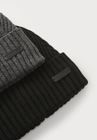 Knit beanies in black and grey color; textured ribbed design with leather logo patches. Soft material; stacked arrangement.