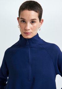 Jeune personne avec les cheveux courts et des boucles d'oreilles en or, portant un haut zippé à col montant bleu marine sur un fond clair et uni.