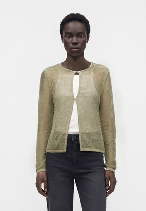 Kvinde iført en lys olivengrøn strikket cardigan over en hvid top og mørke jeans, stående mod en ensfarvet lys baggrund.