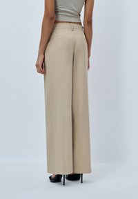 Beige leveled trousers made of smooth fabric, featuring a structured waistband, back pockets, and paired with black high-heeled shoes.