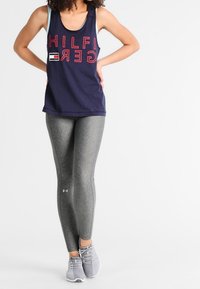 Canotta senza maniche blu navy con testo e logo rossi, abbinata a leggings grigi con texture. Il modello indossa scarpe da ginnastica grigie.