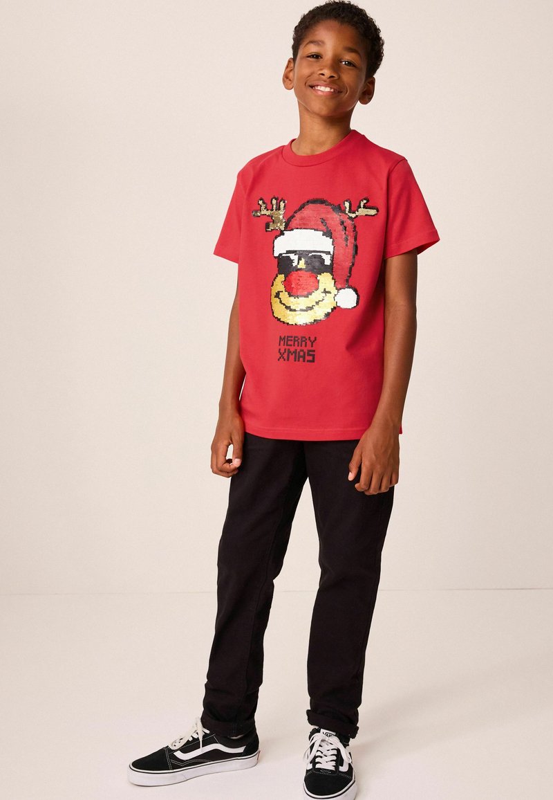 T-shirt en coton rouge avec un graphique d'un renne souriant portant des bois et un bonnet de Santa, texte "JOYEUX NOËL" en dessous en noir. Pantalons noirs et baskets.