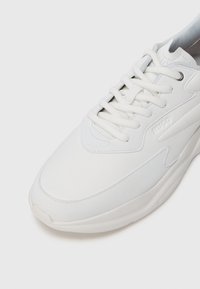 Hvit snøring sneaker med teksturerte paneler og tykk såle, med det pregete merkenavnet "HUGO" på siden.