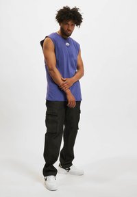 Purple sleeveless t-shirt with a logo on the chest, paired with black cargo pants featuring side pockets and white sneakers.
