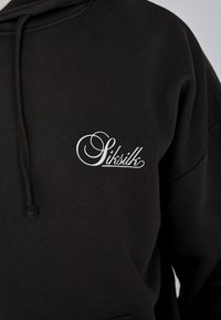 Sudadera negra de tejido suave, con un logo blanco bordado en el área del pecho izquierdo y detalles de cordón en la capucha.