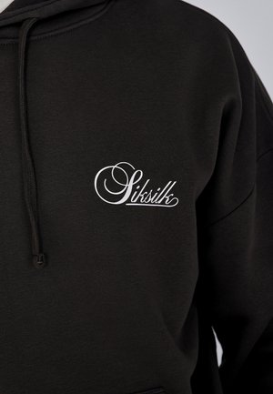 Crna dukserica s kapuljačom, napravljena od mekog materijala, s bijelim izvezenim logom na lijevoj strani grudi i vezicama na kapuljači.