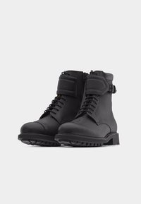 Paire de bottes de combat en cuir noir à lacets, avec fermeture éclair latérale et sangles rembourrées sur fond blanc.
