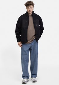 Jachetă din polar neagră cu fermoar frontal, două buzunare pe piept și o etichetă cu logo. Acoperită de un pulover bej și jeans albastri largi.