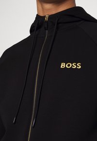 Svart hoodie av mjukt tyg med guldfärgad dragkedja och broderad "BOSS"-logotyp i guld på vänstra bröstet.