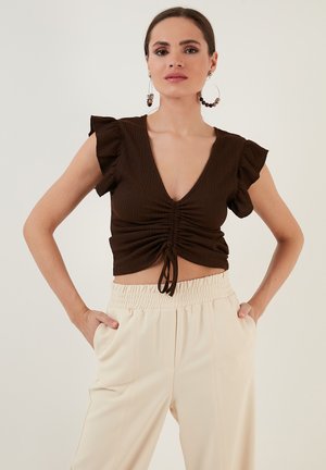 Braun gerippter Crop-Top mit kurzen, schwingenden Ärmeln, tiefem V-Ausschnitt, gerafftem Vorderteil mit Bindeband, kombiniert mit cremefarbenen Hose mit elastischem Bund.
