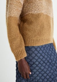 Stickad tröja med en gradientdesign i ljusbrunt och beige, med en texturerad yta. Matchad med en blå texturerad kjol.