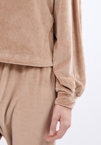 Beżowy, prążkowany top z weluru z okrągłym dołem i luźnymi rękawami. Miękka tekstura z marszczonymi mankietami. Dopasowany w okolicy talii, swobodny krój.