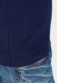 Temno modra bombažna majica s gladko teksturo, z ravnim robom, v kombinaciji s svetlo modrimi jeans hlačami z kontrastnim šivanjem.