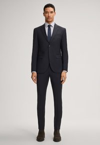 Marineblauer Anzug mit schlanker Passform, einreihigem Blazer, Reverskragen und glatten Fronthosen. Kombiniert mit einem hellen Hemd und einer dunklen Krawatte. Braune Stiefel.