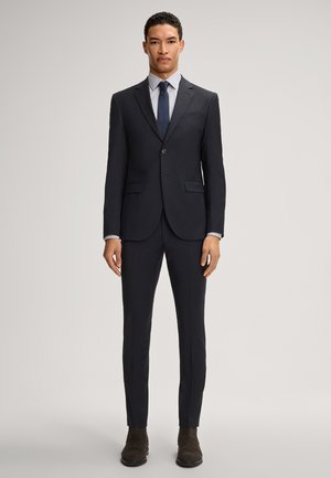 Marineblauer Anzug mit schlanker Passform, einreihigem Blazer, Reverskragen und glatten Fronthosen. Kombiniert mit einem hellen Hemd und einer dunklen Krawatte. Braune Stiefel.