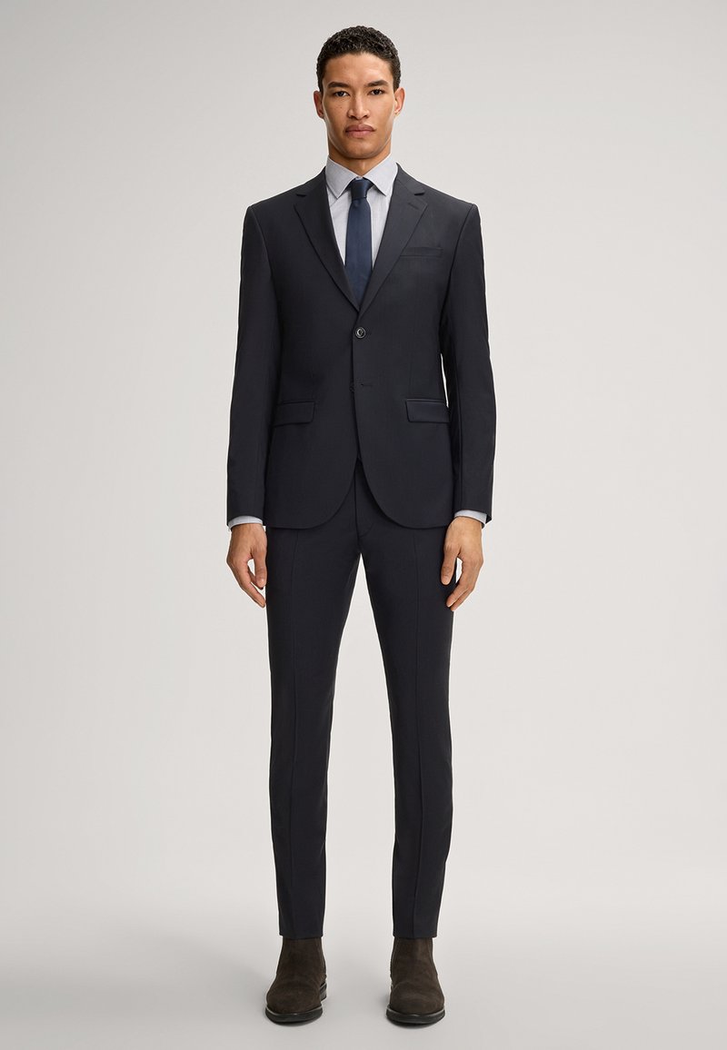 Marineblauer Anzug mit schlanker Passform, einreihigem Blazer, Reverskragen und glatten Fronthosen. Kombiniert mit einem hellen Hemd und einer dunklen Krawatte. Braune Stiefel.