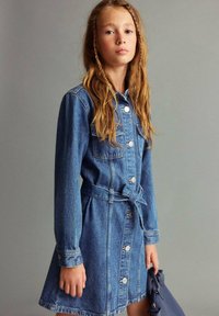 Jeans haljina s dugim rukavima, gumba sprijeda i detaljem oko struka. Svijetloplava boja, džepovi na prsima i blago rašireni kroj suknje.