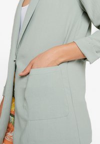 Vaaleanvihreä bleiseri, jossa on yksittäinen nappi, pehmeä kangas ja etutasku. Hihat on käännetty, mikä paljastaa kuvioidun sisäpuolen.