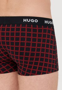 Boxer ajustados negros con un patrón repetido de "HUGO" en rojo, con una cinturilla elástica negra que muestra "HUGO" en letras blancas.