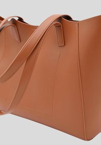 Sac fourre-tout en cuir tan avec deux longues poignées, texture lisse et design minimaliste. Présente une couture verticale sur un côté.