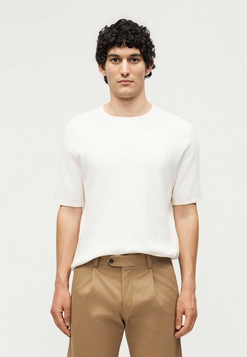 Άντρας με σγουρά μαλλιά που φοράει υφασμάτινο άσπρο πουκάμισο με κοντά μανίκια και μπεζ ψηλόμεσα παντελόνια σε απλό ανοιχτό φόντο.